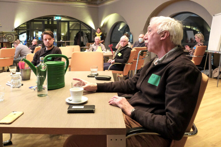 Een man zit aan tafel en praat naar links toe.